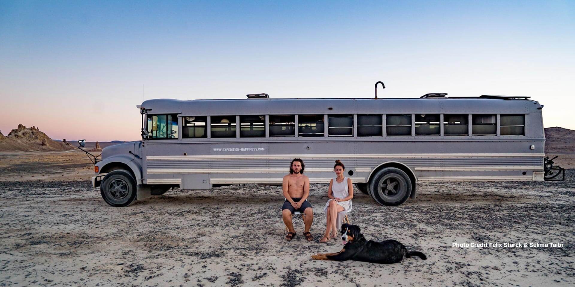 Two people with their dog sitting in front of a bus in the dessert